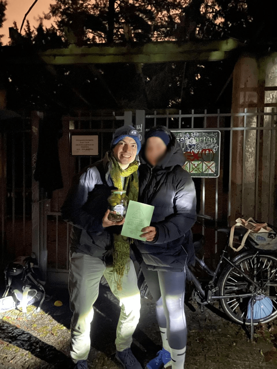 Zwei Läufer mit Stirnlampen vor einem Parktor, einer hält ein Glas Gurken und eine Karte in der Hand